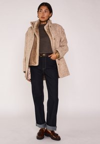Cappotto in misto lana beige con un interno morbido e testurizzato, jeans scuri con risvolti, e mocassini marroni con nappine. Orecchini a cerchio dorati completano il look.