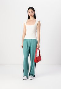 Hvit ribbet tanktop, teal løse joggebukser med sidestriper, hvite sneakers og en liten rød skulderveske. Enkel hvit bakgrunn.