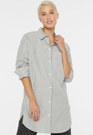 Junge Person mit kurzen blonden Haaren, die ein langes, weißes Hemd mit dünnen vertikalen schwarzen Streifen und hochgekrempelten Ärmeln trägt.