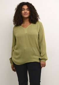 Blusa da donna verde oliva a maniche lunghe con scollatura a V e una sottile trama. Presenta polsini elastici e una vestibilità morbida. Indossata con pantaloni scuri.