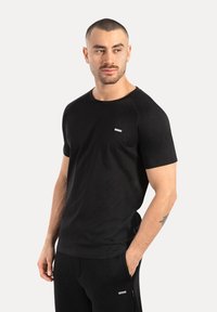 Man met kort haar en baard, gekleed in een zwart T-shirt met korte mouwen en zwarte broek, staande met één hand in zijn zak, kijkend naar de zijkant.