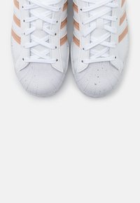 Vita atletiska skor med tre persikofärgade ränder, texturerad gummitå, och platta vita skosnören mot en enfärgad bakgrund.