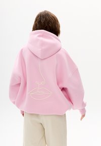 Oversized Hoodie in Pink aus weichem Stoff, mit einer schlichten Linienzeichnung von Lippen und Nase auf der Rückseite. Weiße Stickerei-Details.