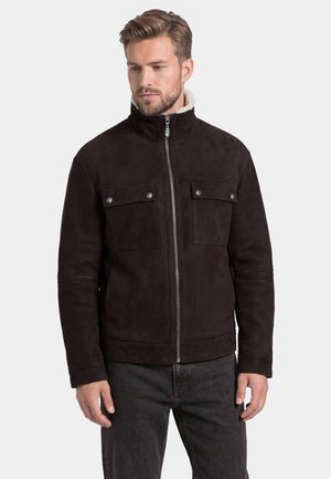 Mann trägt eine dunkelbraune Jacke mit Reißverschluss, zwei mit Knöpfen verschlossene Brusttaschen und eine hell gefütterte Innenseite am Kragen.
