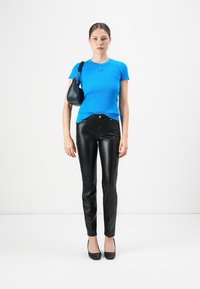 Vrouw staat rechtop en draagt een heldere blauwe korte mouwen shirt, zwarte leren broek, zwarte hakken en een kleine zwarte schoudertas.