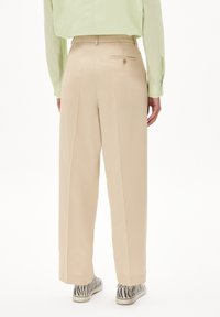 Beige wijde broek met een gladde textuur, voorzien van een achterzak met een knoop. Gekozen met een lichtgroen shirt en gestreepte schoenen.