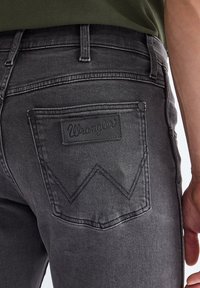 Achteraanzicht van een donkergrijze Wrangler spijkerbroekzak met logopatch en stikseldetails, gedragen door een persoon in een groen shirt.