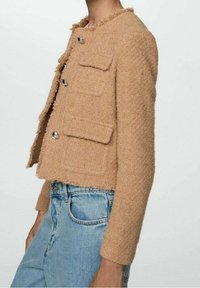 Beige zkrácená bunda vyrobená z texturované tkaniny, s čtyřmi předními kapsami, stříbrnými knoflíky a třásněmi podél výstřihu a lemu.