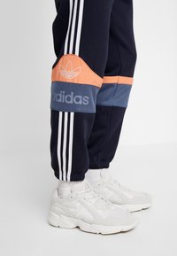 Mörkblå joggingbyxor med vita ränder, en orange detalj och en blå panel med varumärke; kombinerat med vita sportskor.