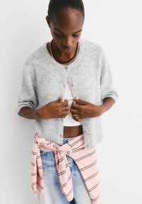 Grau, fusseliger Cardigan mit kurzen Ärmeln, Knopfschluss und einem rosa gestreiften Shirt, das um die Taille gebunden ist, über hellblauen Jeans.
