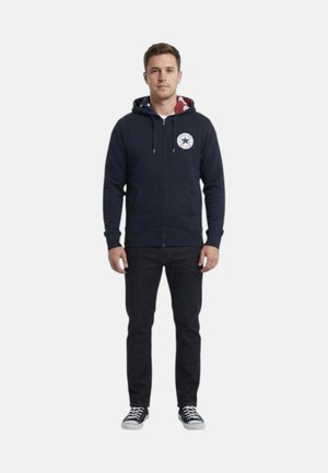 Uomo in piedi di fronte, indossa una felpa con cappuccio blu navy con zip e logo a stella, jeans neri e sneakers nere con lacci bianchi.