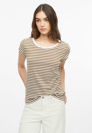 Jeune femme aux longs cheveux bruns portant un t-shirt à manches courtes rayé marron et blanc et un pantalon blanc, debout les mains derrière le dos.