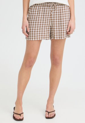 Nohy v hnedom a bielom károvanom šortkách so šnúrkou a tmavohnedých žabkách, stojace na jednoliatom pozadí.