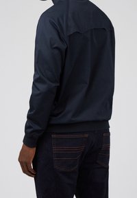 Navy blue lightweight jacket with a high collar, elastic cuffs, and hem. Features contrasting stitching on dark denim jeans.