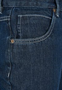 Mørkeblå denim jeans med en synlig baglomme, hvide stikninger og en metalknapdetalje ved lommeåbningen.