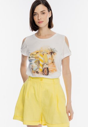 Kobieta ubrana w białą koszulkę z odkrytymi ramionami, z nadrukiem vintage skutera i palmy, zestawioną z żółtymi szortami z wysokim stanem.