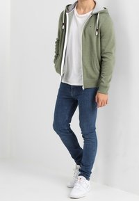 Grüne Zip-Hoodie mit grauem Innenfutter, kombiniert mit einem weißen T-Shirt und blauen Skinny Jeans. Abgerundet mit weißen Sneakers. Einfaches Design, lässiger Look.