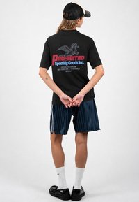 Άτομο που φοράει μαύρο καπέλο, μαύρο T-shirt με λογότυπο Πήγασου που γράφει "Prohibited Sporting Goods Inc.", ναυτικό ριγέ σορτς, λευκές κάλτσες και μαύρα παπούτσια.