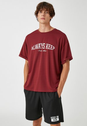 Camiseta de manga corta color marrón burdeos con texto blanco en el frente, combinada con pantalones cortos negros que tienen un acento de patrón blanco en la pierna.