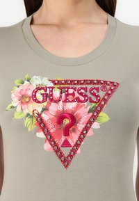 Pilkas prigludęs marškinėliai su dideliu gėlių raštu, turinčiu trikampį, papuoštą raudonu „GUESS“ logotipu ir deimantais, su tekstūruotu paviršiumi.