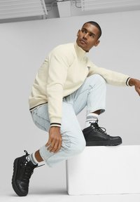 Kremowa bluza na zamek błyskawiczny z czarnymi akcentami, jasnoniebieskie dżinsy z podwiniętymi nogawkami oraz czarne trampki za kostkę z białymi i czarnymi paskami skarpet.