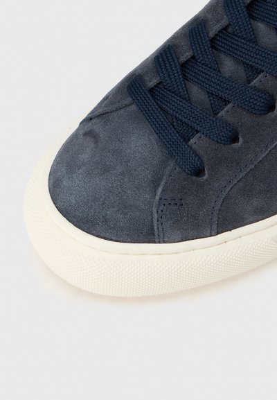 Ναυτικό σουέντ αθλητικό παπούτσι με υφή στο πάνω μέρος, διαθέτει κορδόνια στο ίδιο χρώμα και λευκή λαστιχένια σόλα, τονίζοντας καθαρές γραμμές και μίνιμαλ σχεδιασμό.