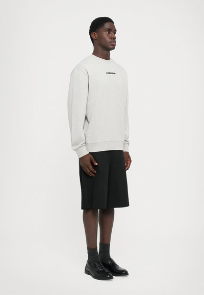 Mann iført lys grå hettegenser med logo, svarte vide shorts til knærne, mørke sokker og svarte lærsko, står mot en hvit bakgrunn.