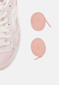 Baskets rose clair en mesh et suède, avec un logo blanc et des lacets plats. Deux lacets ronds roses sont présentés à côté.