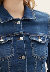 Jeansjacka i mörkblått med metalliska silverknappar, två bröstfickor och en spetsig krage. Har kontrasterande sömmar.