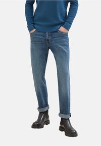 Suéter azul de cuello redondo, jeans azul claro con puños enrollados y botas negras hasta el tobillo. El denim tiene un ligero desgaste; el suéter tiene un tejido texturizado.
