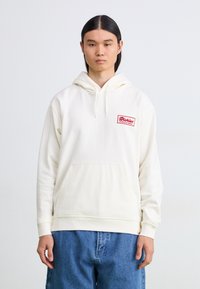 Off-white hettegenser med lomme på forsiden, ribbede mansjetter og en rød brodert logo-patch på venstre bryst. Denimbukser.