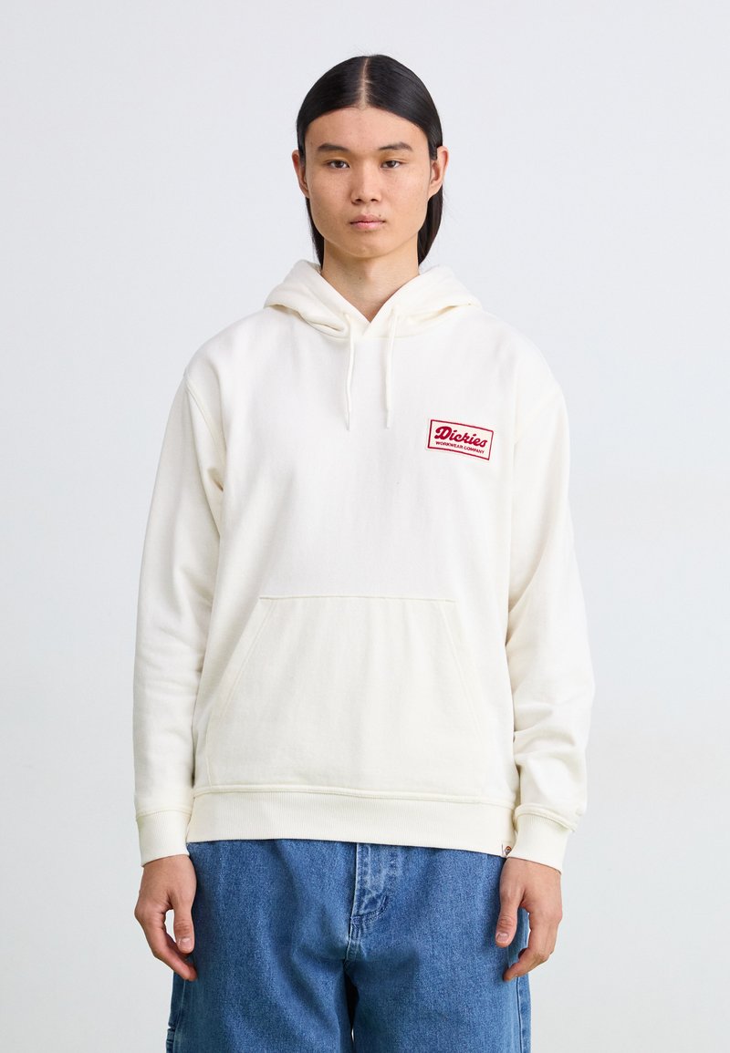 Off-white hettegenser med lomme på forsiden, ribbede mansjetter og en rød brodert logo-patch på venstre bryst. Denimbukser.