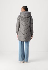 Manteau doudoune gris avec un design en chevrons, doté d'une capuche et de poignets côtelés, porté sur un jean large bleu et des baskets blanches.