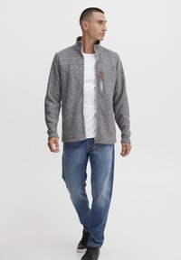 Giacca a maglia grigia con colletto alto, chiusura con zip e dettagli arancioni. Abbinata a jeans in denim azzurro chiaro e sneakers nere.