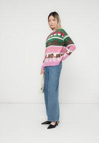 Strikket sweater med grønne, pink og hvide julemønstre, der har snefnug og gaver. Kombineret med lyseblå brede jeans og sorte sko.