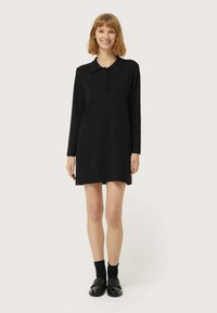 Vestido negro de manga larga con cuello, botones en el frente y sutilezas en los laterales. Lleva calcetas negras y mocasines negros brillantes.