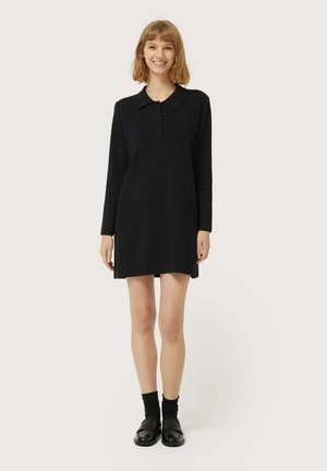 Vestido negro de manga larga con cuello, botones en el frente y sutilezas en los laterales. Lleva calcetas negras y mocasines negros brillantes.