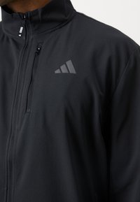 Černá sportovní bunda s vysokým límcem, hladkým povrchem, plným zipem a malým reflexním trojúhelníkovým logem na levé hrudi.