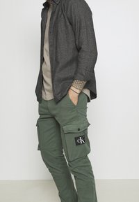 Chemise texturée grise portée par-dessus un t-shirt beige, associée à un pantalon cargo vert avec poches latérales et un logo en patch noir.