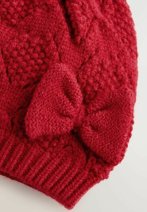 Rode gebreide muts met een textuurpatroon, voorzien van een grote strikaccent. Het materiaal lijkt zacht en chunky met ribbelranden.