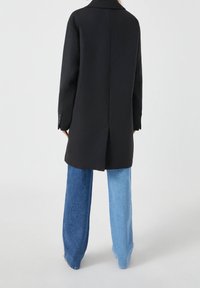 Černý oversized kabát s jedním rozparkem vzadu, doplněný širokými džíny v kontrastních světlých a tmavých modrých odstínech.
