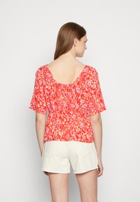 Blusa floral vermelha com mangas curtas, decote quadrado nas costas e elástico na cintura, combinada com calções cremes.