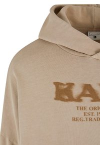 Haut à capuche beige en tissu doux avec un grand texte graphique marron pâle "KANK" et de petits détails en dessous, doté d'une capuche avec cordon.