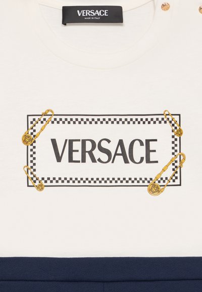 Camisa de algodón blanca con un borde a cuadros en blanco y negro y alfileres de seguridad dorados. La palabra "VERSACE" está impresa en negro de manera prominente.
