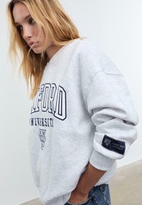 Mlada žena s plavom kosom nosi svijetlosivi oversized duksericu Oxford University i plave traperice, stoji s rukama u džepovima.