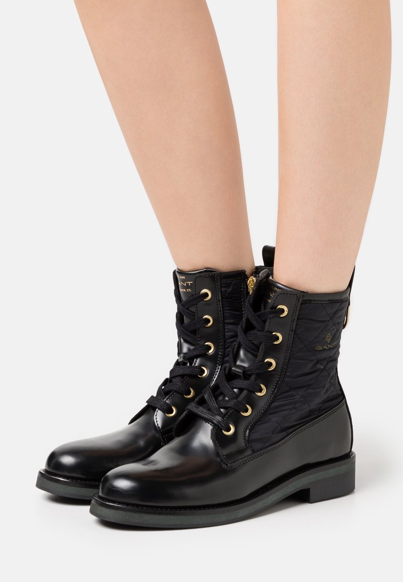 Bottines montantes en cuir noir et tissu matelassé avec lacets, œillets dorés et fermetures éclair, portées sur jambes nues sur fond blanc.