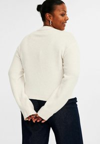 Leende kvinna med mörkt hår i en knut, iförd silverörhängen i form av ringar, en krämfärgad tröja och mörka jeans, med händerna knäppta bakom ryggen.
