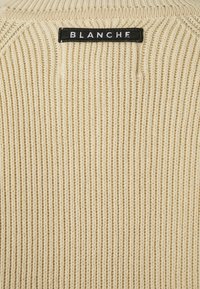 Lys beige strikkegenser med ribbestruktur, rund hals og en svart etikett med ordet "BLANCHE" brodert bak. 