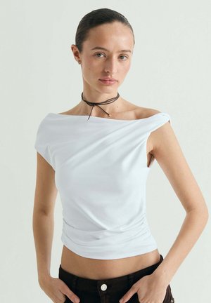 Mujer con el cabello peinado hacia atrás lleva una blusa blanca sin hombros, pantalones negros y un collar choker negro y delgado, posando con las manos en las caderas.