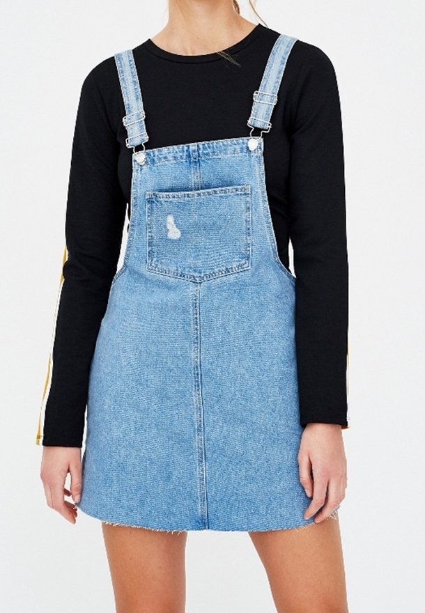 Femme portant une robe salopette en denim bleu clair par-dessus un chemisier noir à manches longues, debout les mains détendues le long du corps.